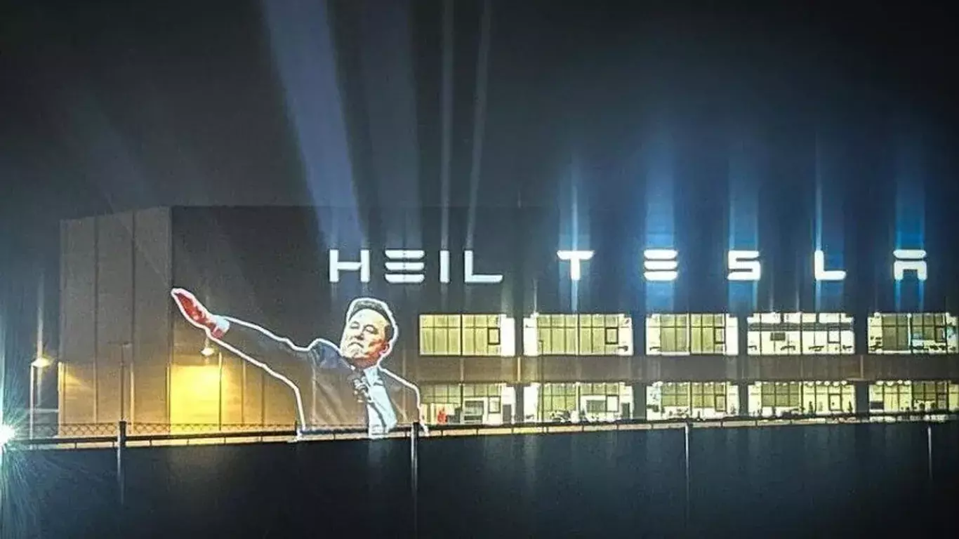 A picture of a tesla factorio with a projection of musk doing a nazi salute and the word "HEIL" before "TESLA".