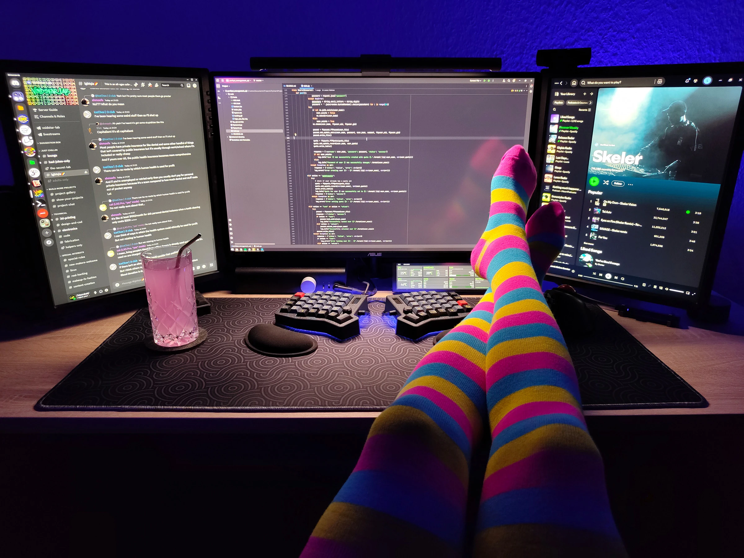 Photo of my desk with three computer monitors on it. The wall behind the monitors is lit up with blue light. My legs in pink, yellow and blue striped thigh highs are resting on the desk next to my keyboard. On the left side of the desk is a glass filled with pink lemonade.