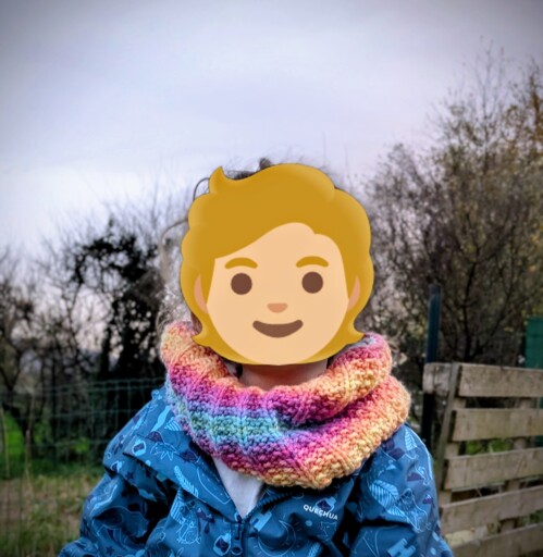 Un portrait d'une petite fille avec un tour de cou tricoté multicolore. La tête est remplacée par un emoji.