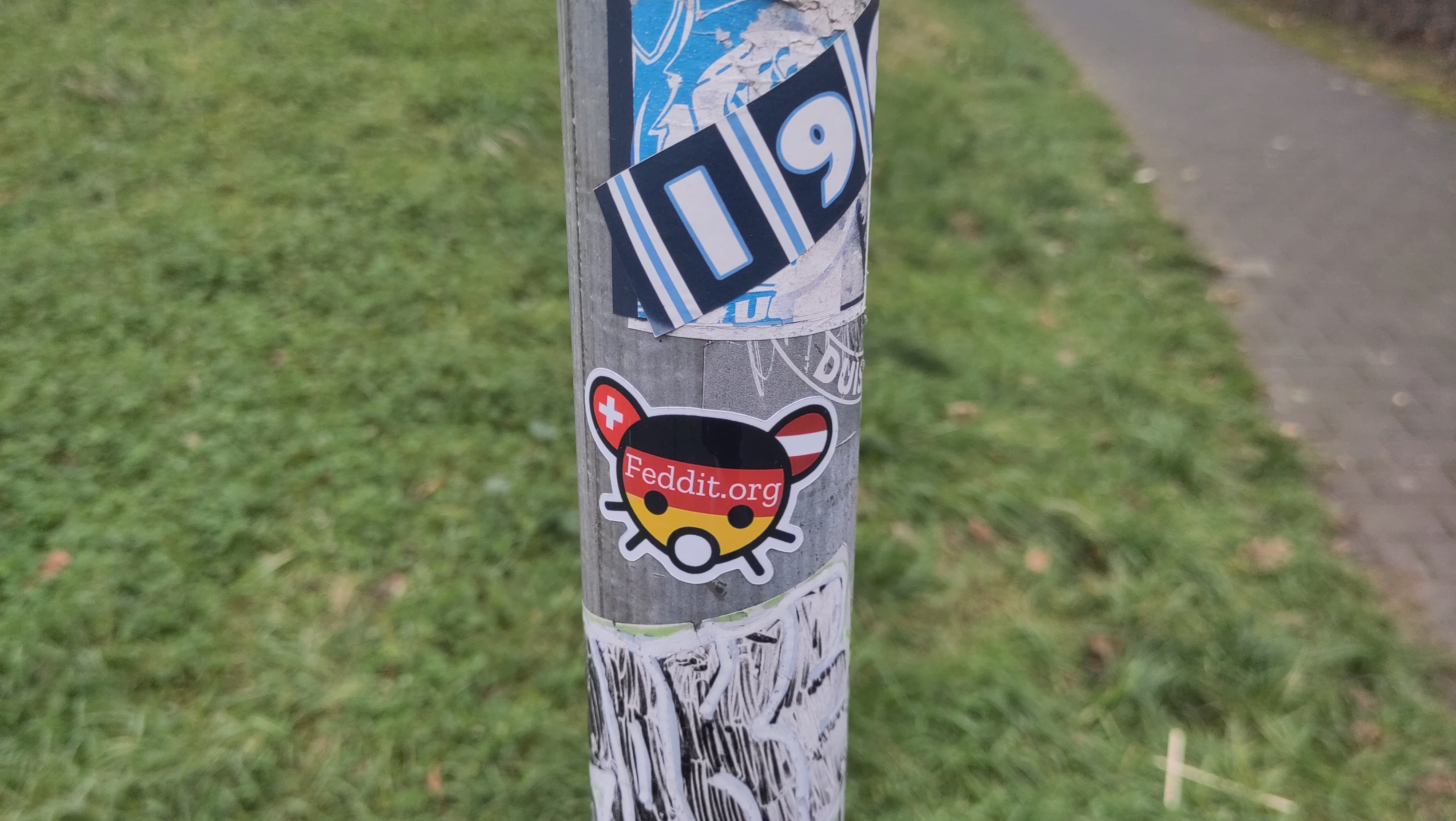 Metallpfahl mit verschiedenen Stickern, mittig ein feddit.org Sticker.