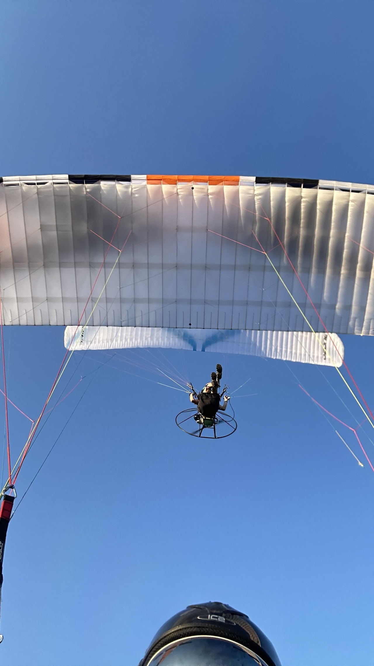 Selfie shot, from paramotor, with helmet and goggles in bottom edge of shot. Above you see the orange, white, and black wing. Above and just back of that you see another paramotor pilot with a blue and white wing.