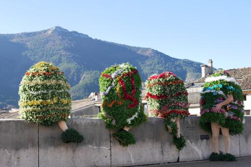 Photographie de quatre personnes déguisées en sapins de Noël, assises au bord d’un pont. On ne voit pas leur tête, seules les jambes (voire les bras) dépassent du « sapin », et ça donne un impression assez bizarre et rigolote. Il y a des toits de maisons et une jolie montagne en arrière-plan.